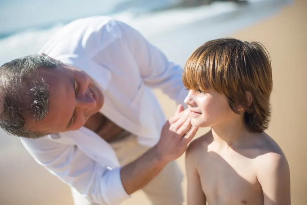 Mains d'un parent appliquant une crème solaire sur un bébé à l'ombre, avec chapeau et bouteille d'eau